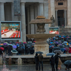 Faithful visitors, holding umbrellas, look at a giant screen showing the start of the papal conclave, under a statue of St. Peter on St. Peter's Square on Tuesday (March 12) at the Vatican. RNS photo by Andrea Sabbadini