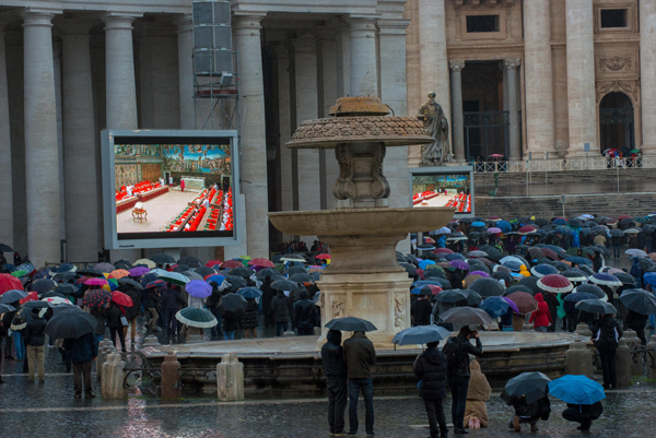 Faithful visitors, holding umbrellas, look at a giant screen showing the start of the papal conclave, under a statue of St. Peter on St. Peter's Square on Tuesday (March 12) at the Vatican. RNS photo by Andrea Sabbadini