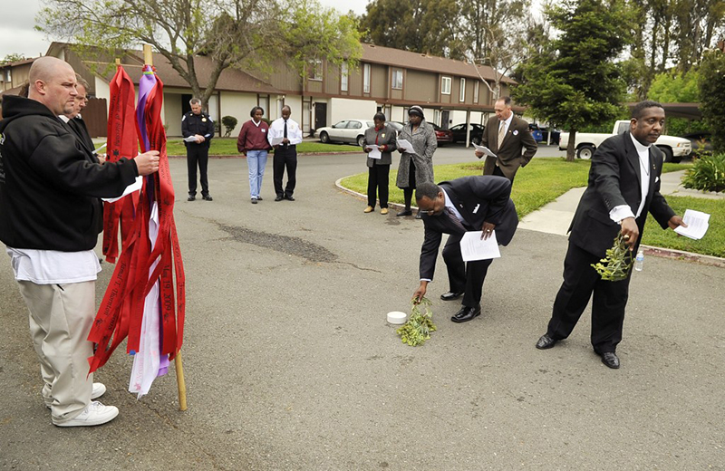 A moment of blessing in Fairfield, Calif. at the Parkway Gardens apartment complex takes place between clergy and law enforcement. Pictured here are, fire chief Vincent Webster, center right, Captain Darrin Moody, in uniform, center left, Brother Ray Courtamathe, reading in a brown jacket, the Rev. Vic Russel, second from right, and the Rev. David Isom, far right. A far left, David Derf and Todd Bertani hold poles that carry names of victims of homicide and violent death. The light pink flags stand for children killed. Photo by Jess Sullivan