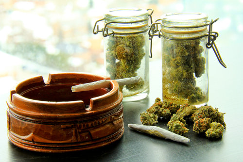 A marijuana joint in an ashtray, with another joint, a pile and two jars full of more marijuana.