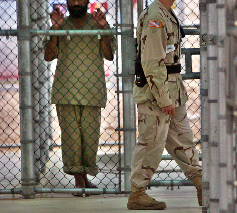 In this photo reviewed by U.S. military officials, a detainee whose name, nationality, and facial identification are not permitted, holds onto a fence as a U.S. military guard walks past the grounds of the maximum security prison at Camp 5 in the Guantanamo Bay U.S. Naval Base on June 26, 2006. Photo courtesy of REUTERS