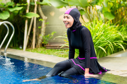 Muslim woman or girl sitting at pool in tropical garden wearing Burkini halal swimwear.