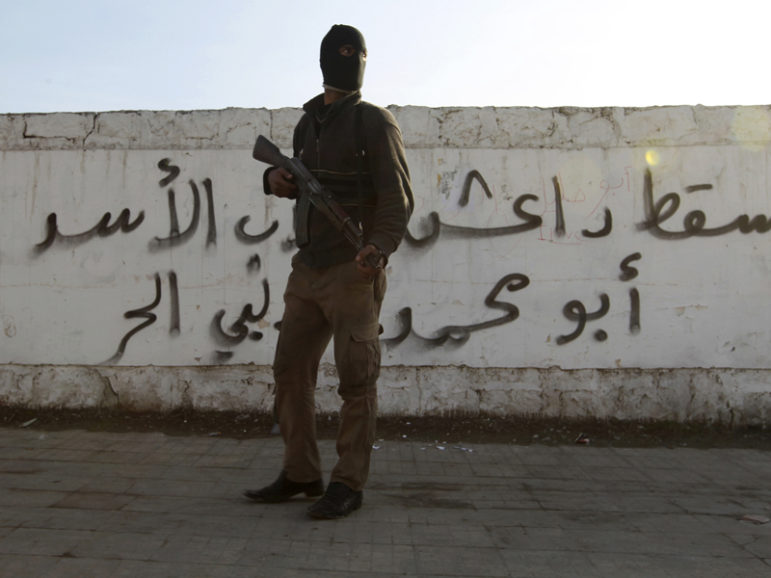 A Free Syrian Army fighter carries his weapon as he stands in front of graffiti that reads  