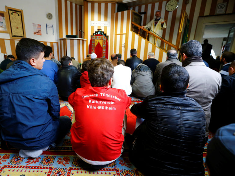 Young Muslims listen to a Turkish imam during Friday prayers at the Turkish Kuba Camii mosque located near a hotel housing refugees in Cologne's district of Kalk, Germany, Oct. 14, 2016. Youth in foreground wears a shirt reading, 