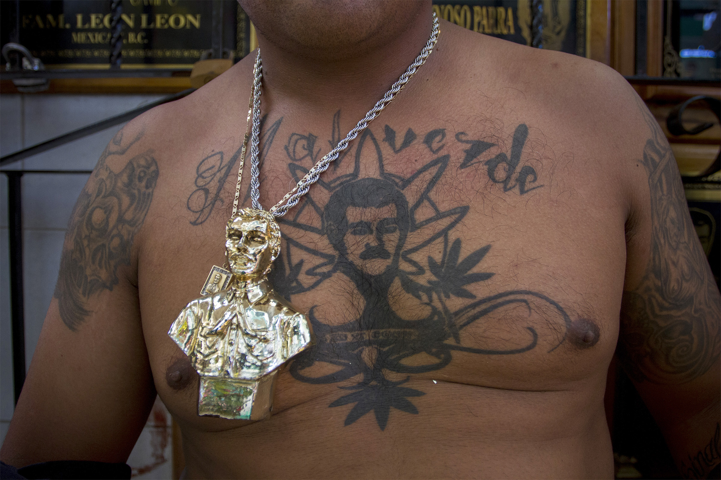 A man wears a chain with a large bust of Jesus Malverde on top of a tattoo
