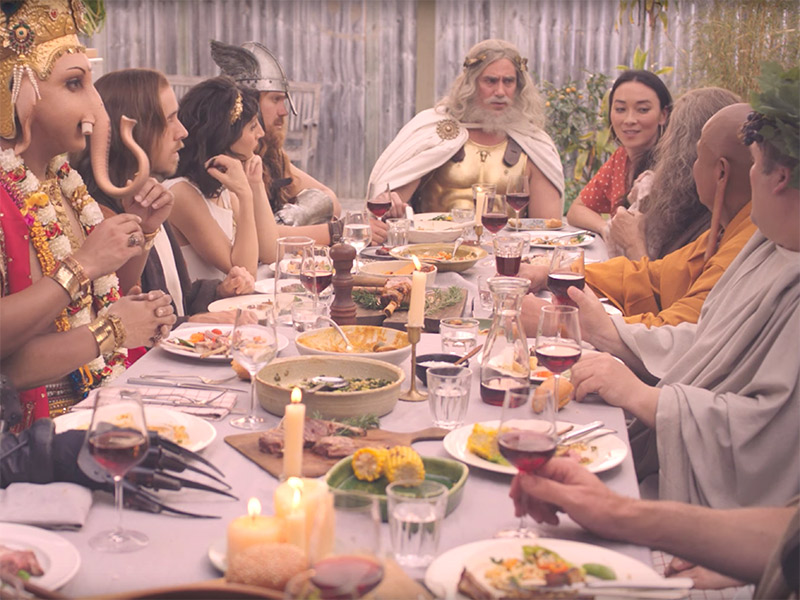 Various gods, goddesses and prophets including Jesus, God, Aphrodite, Zeus, Buddha, Ganesh and Moses sit at a long dining table to enjoy a spring barbecue of lamb. 