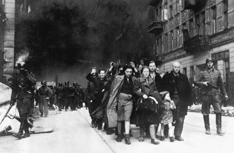 Jewish civilians are led by German Nazi troops to the assembly point for deportation. Picture taken at Nowolipie street, near the intersection with Smocza duing Warsaw Ghetto Uprising in April 1943. Housing blocks burn in the background. Photo courtesy of NARA/Creative Commons