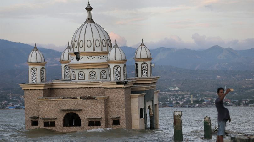 A man selfies with the background of a mosque that is isolated by water after its bridge was broken due to the massive earthquake and tsunami in Palu, Central Sulawesi, Indonesia, Saturday, Oct. 6, 2018. A 7.5 magnitude earthquake rocked the city on Sept. 28, triggering a tsunami and mud slides that killed a large number of people and displaced tens of thousands others. (AP Photo/Tatan Syuflana)