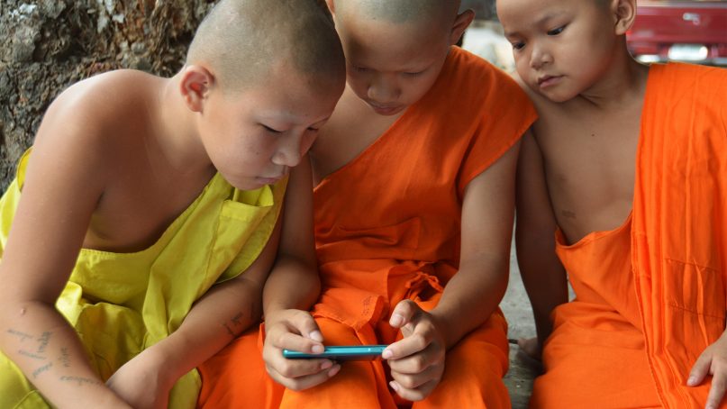 Novice monks look at a smartphone. Buddhism has reached many adherents through social media and online videos. Photo courtesy of Creative Commons