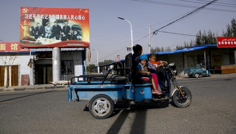 FILE - In this Sept. 20, 2018, file photo, an Uighur woman shuttles school children on an electric scooter as they ride past a propaganda poster showing China's President Xi Jinping joining hands with a group of Uighur elders in Hotan, in western China's Xinjiang region. Under Chinese President Xi Jinping, the Uighur homeland has been blanketed with stifling surveillance, from armed checkpoints on street corners to facial-recognition-equipped CCTV cameras steadily surveying passers-by. Now, Uighurs say, they must live under the watchful eye of the ruling Communist Party even inside their own homes. (AP Photo/Andy Wong, File)