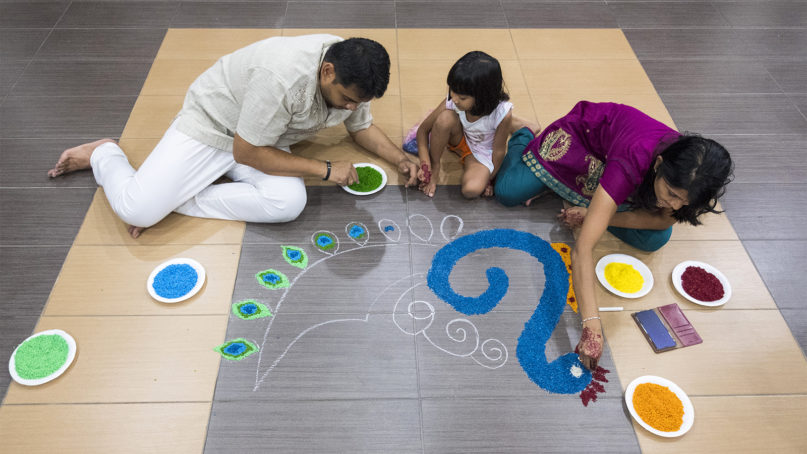 Krishna Shanmugam, left, makes a kolam in front of his parents house with his daughter Mageswari, 4 and his wife, Jasmira, on Oct. 26, 2019, the eve of Deepavali. The kolam, an intricate Indian folk art design made from colorful rice or rice flour, usually decorates the entrance of the house on Deepavali. It is a display meant to welcome prosperity and blessings. This year the Shanmugams chose to make a peacock kolam, an animal which is considered auspicious to Hindus, symbolizing kindness, compassion, safeguarding and knowledge. RNS photo by Alexandra Radu