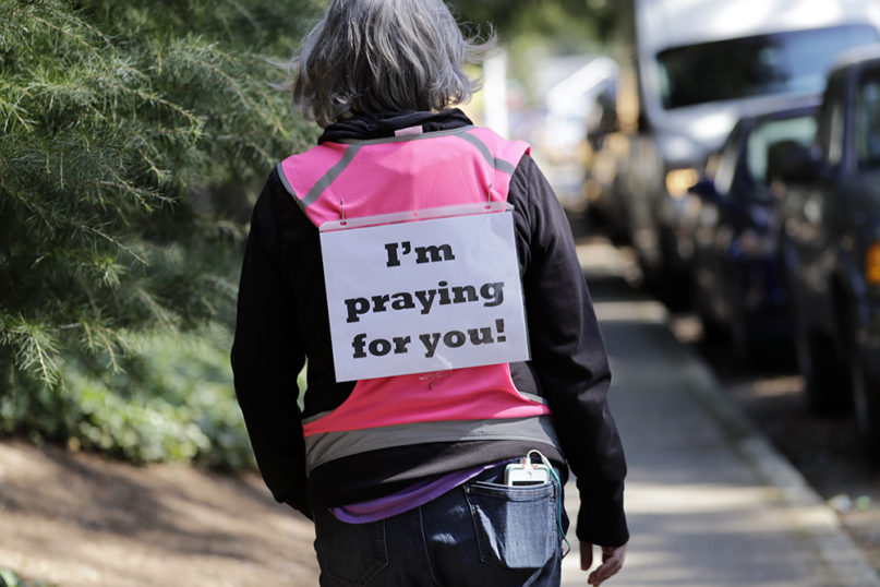 Lois Casimes wears a sign in support of residents and health care workers as she walks past the Life Care Center, where at least 30 coronavirus deaths have been linked to the facility, on March 18, 2020, in Kirkland, Washington. Casimes, from nearby Bothell, Washington, wanted to take the time to walk in the neighborhood and pray for people at the facility. (AP Photo/Elaine Thompson)