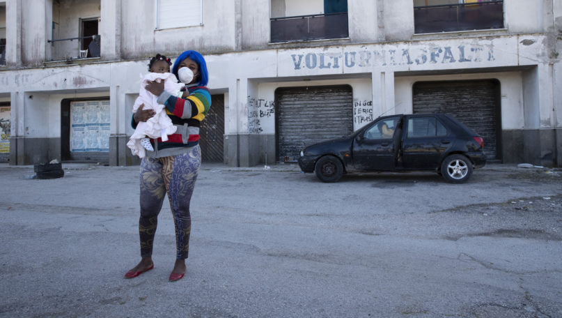 FILE -- In this photo taken on Monday, April 27, 2020, a woman wearing a sanitary mask to protect against COVID-19 holds her daughter as she walks past a building in Castel Volturno, near Naples, Southern Italy. They are known as “the invisibles,” the undocumented African migrants who, even before the coronavirus outbreak plunged Italy into crisis, barely scraped by as day laborers, prostitutes and seasonal farm hands. Pope Francis is calling for migrant farm workers to be treated with dignity, issuing an appeal as Italy weighs whether to legalize undocumented agricultural workers amid a shortage of seasonal farm labor due to the coronavirus emergency. (AP Photo/Alessandra Tarantino)