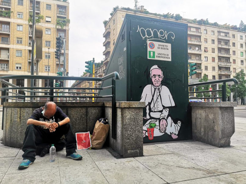 A man sits by a pop art graffiti titled “Caritas,” which depicts Pope Francis begging, by aleXsandro Palombo, in Milan. Photo courtesy of aleXsandro Palombo