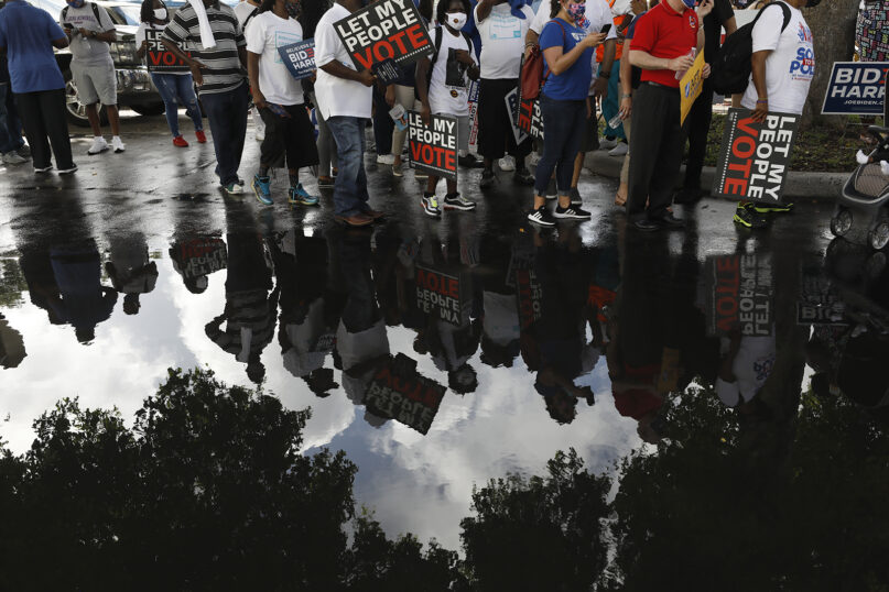 People carrying signs supporting voting rights are reflected in a puddle as they arrive at an early voting center at Model City Branch Library, as part of a 