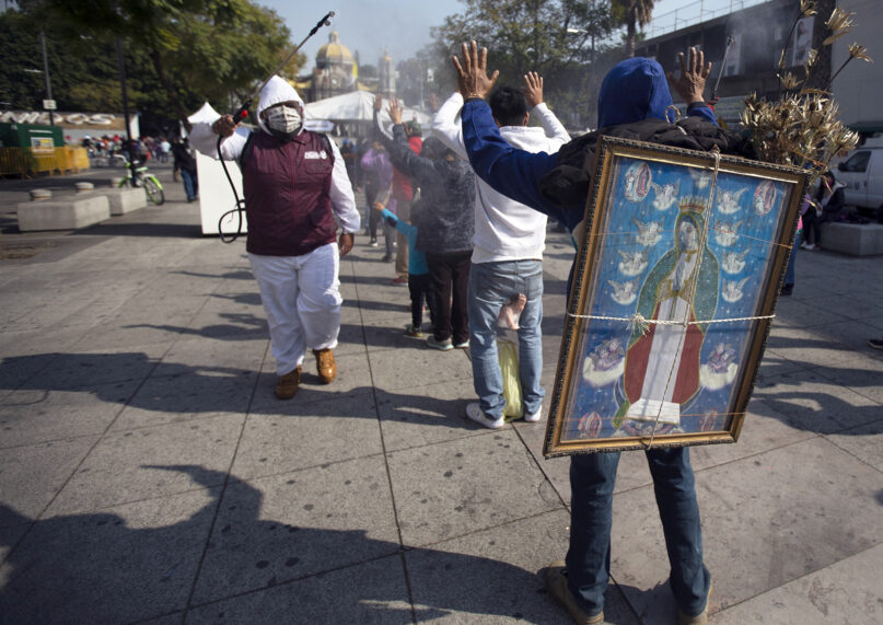 A health worker disinfects pilgrims on their way to the Basilica of the Virgin of Guadalupe to curb the spread of the new coronavirus, as Alejandro Castillo carries an image of the Virgin on his back Dec. 9, 2020, in Mexico City. Castillo departed his home in San Gregorio Atzompa, Puebla state, along with his family to make the pilgrimage to the basilica on Dec. 6, and made it to the temple a day before it closed to the public due to the COVID-19 pandemic. (AP Photo/Marco Ugarte)