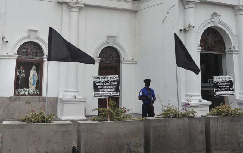 A policeman stands guard as black flags are seen in front of St. Anthoney's Church, in Colombo, Sri Lanka, Saturday, Aug. 21, 2021. Sri Lankan Catholics hoisted black flags in churches and at homes on Saturday protesting against what they call government’s inaction to find  the true conspirators in the Easter Sunday blasts of 2019 which killed 269 people. (AP Photo/Eranga Jayawardena)