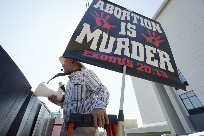 Anti-abortion activist Doug Lane uses a ladder to peer over the covered fencing as he calls out to patients entering the Jackson Women’s Health Organization clinic in Jackson, Mississippi, moments after the U.S. Supreme Court ruling overturning Roe v. Wade was issued, June 24, 2022. The clinic is the only facility that performs abortions in the state. (AP Photo/Rogelio V. Solis)