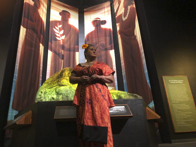 Hinaleimoana Wong-Kalu, one of the curators of the new Kapaemahu exhibit at Bishop Museum, poses for a photo in Honolulu on Thursday, June 16, 2022 in front of pictures of four healers who visited Hawaii from Tahiti more than 500 years ago. The exhibit draws attention to the stories of the healers, who were 
