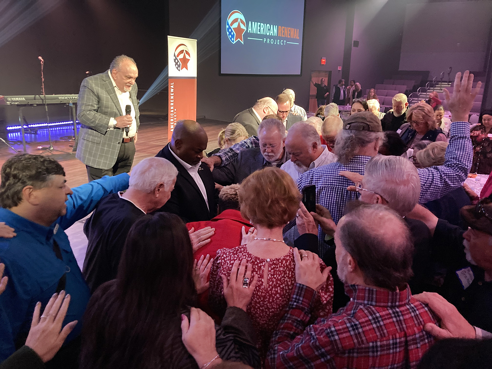 Pastors lay hands and pray for Lt. Gov. Mark Robinson of North Carolina, center left, at a pastors luncheon hosted by the American Renewal Project in Raleigh, North Carolina, on Oct. 10, 2022. The Rev. Gary Miller, on stage, leads the prayer. RNS Photo by Yonat Shimron