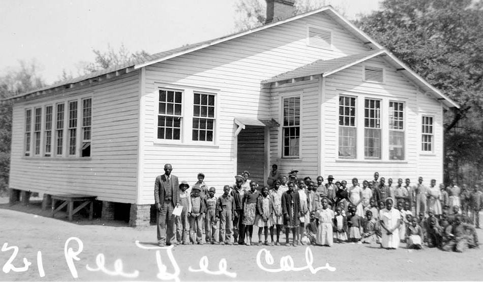 People pose outside the Pee Dee Rosenwald School in Marion County, South Carolina, c. 1935. The Pee Dee Rosenwald School was built in 1922-1923 with two classrooms and two cloakrooms. A chalkboard was located on the partition between the two rooms. Students' desks faced the chalkboard with the teachers' desks located off to the side. The wall between the classrooms could be moved to open a large space in the school. Photo from the South Carolina Department of Archives and History/Creative Commons