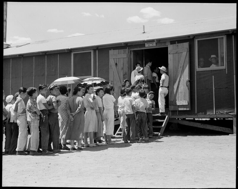 Part of a line waiting for lunch outside the mess hall at the Manzanar War Relocation Center in Manzanar, Calif., on July 1, 1942. Photo by Dorothea Lange/NARA/Creative Commons