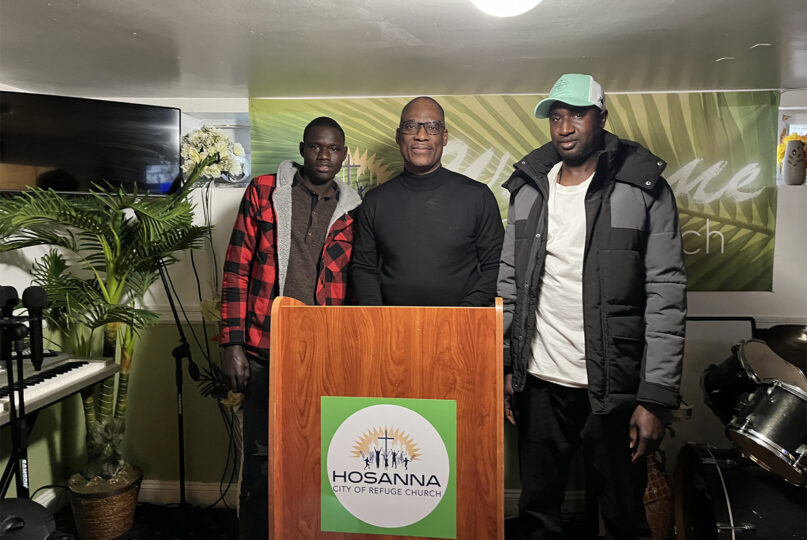 The Rev. Phillip Falayi, center, poses with Ndao Nbaye, left, and Cheikh Dramé, two migrants who are currently sheltering at the Hosanna City of Refuge Church in Queens, New York. (RNS photo/Fiona André)