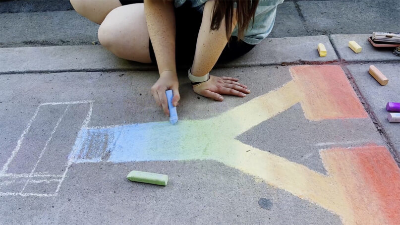 An individual chalks a Y with rainbow colors ahead of a Pride Night in Provo, Utah. (Image courtesy of 