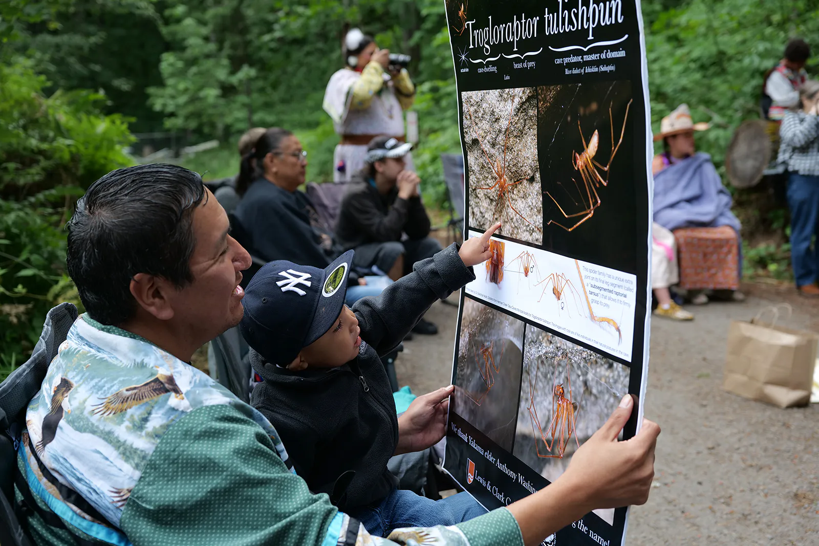 Newly elected Yakama tribal councilman John Washines holds his 2-year-old grandson, Jakoby Kenoras, while they look at a poster featuring pictures of the newly discovered Trogloraptor tulishpun during the spider’s naming ceremony on June 10, 2026, at Herman Creek Campground east of Portland, Oregon. (Photo by Lyric Aquino, Underscore Native News + Report for America)